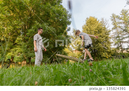 A father watches his child play on a seesaw at the playground, filled with laughter and joy. They are surrounded by trees and vibrant grass, enjoying a beautiful day outdoors 135824391