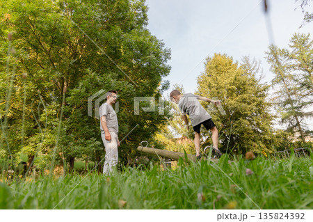 A father watches with joy as his child playfully balances on a wooden beam at a playground. They are immersed in the beauty of nature during a warm afternoon 135824392