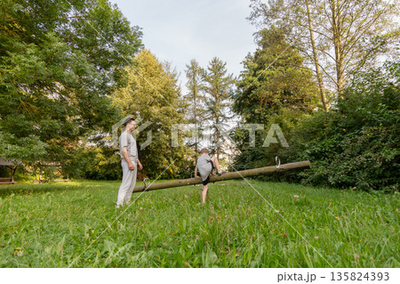 A father and his young child engage in playful activities at a playground, surrounded by vibrant greenery and trees, creating joyful memories in nature during the day A father and his young child engage in playful activities at a playground, surrounded by vibrant greenery and trees, creating joyful memories in nature during the day 135824393