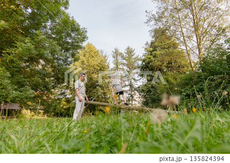 A father watches his child play on a balance beam in a lush, green playground surrounded by trees. They are enjoying a sunny, relaxed day outdoors together A father watches his child play on a balance beam in a lush, green playground surrounded by trees. They are enjoying a sunny, relaxed day outdoors together 135824394