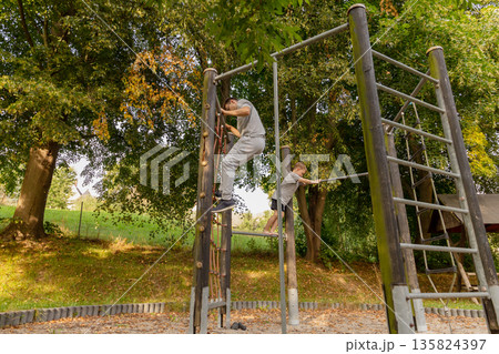 A father and his child are joyfully spending time together at a playground, climbing on the equipment while surrounded by lush greenery on a sunny day A father and his child are joyfully spending time together at a playground, climbing on the equipment while surrounded by lush greenery on a sunny day 135824397