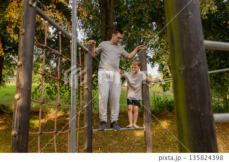 A father helps his child navigate a playground structure, fostering connection and playfulness amidst lush greenery on a sunny day. Their joy reflects the beauty of family bonds A father helps his child navigate a playground structure, fostering connection and playfulness amidst lush greenery on a sunny day. Their joy reflects the beauty of family bonds 135824398