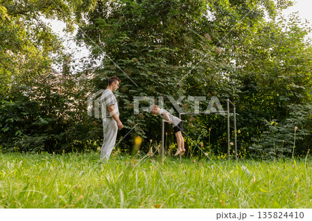 A father encourages his child as they play together on a playground. The child is attempting to perform a gymnastic move while surrounded by lush greenery A father encourages his child as they play together on a playground. The child is attempting to perform a gymnastic move while surrounded by lush greenery 135824410