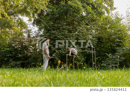 Father watches with joy as his child climbs on playground equipment, surrounded by lush greenery, creating cherished memories in a peaceful outdoor setting 135824411