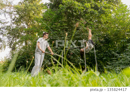 A father guides his child as they engage in playful activities at a playground. The child attempts to perform a handstand while surrounded by vibrant trees and grass A father guides his child as they engage in playful activities at a playground. The child attempts to perform a handstand while surrounded by vibrant trees and grass 135824417