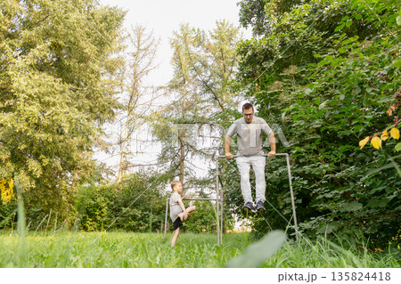 Father and child engage in fun activities at a playground, with the child watching as the father performs exercises. They are surrounded by greenery under a sunny sky Father and child engage in fun activities at a playground, with the child watching as the father performs exercises. They are surrounded by greenery under a sunny sky 135824418