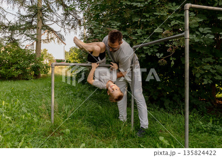 Father and child are enjoying playful moments at a playground, with the child hanging upside down from the bars. Surrounded by lush greenery, they share joy and bonding 135824422