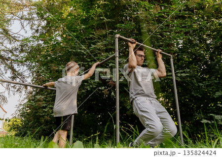 A father and his child are sharing joyful time at a playground, engaging in playful exercises like pull-ups amidst lush greenery during a sunny day A father and his child are sharing joyful time at a playground, engaging in playful exercises like pull-ups amidst lush greenery during a sunny day 135824424