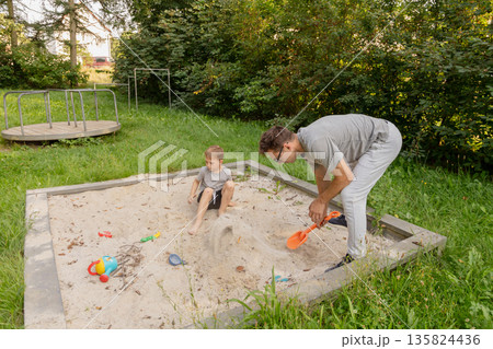 A father and his young child play together in a sandbox, creating shapes and using colorful toys. They are enjoying a sunny day in a park filled with trees and vibrant nature A father and his young child play together in a sandbox, creating shapes and using colorful toys. They are enjoying a sunny day in a park filled with trees and vibrant nature 135824436