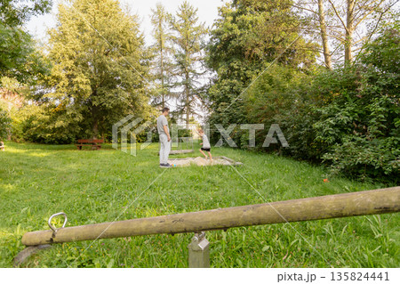 A father plays with his child at a park playground, surrounded by lush trees and grass. They share laughter and joy while enjoying the outdoors on a bright day A father plays with his child at a park playground, surrounded by lush trees and grass. They share laughter and joy while enjoying the outdoors on a bright day 135824441
