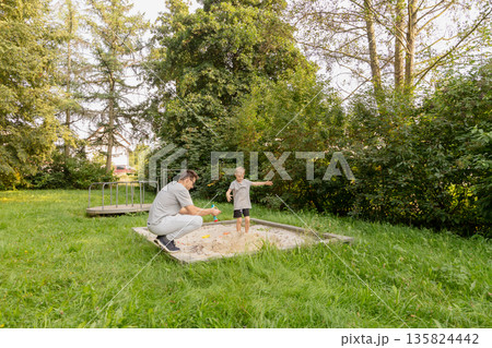 A father and his child are having fun at a playground, engaging in activities at a sandpit surrounded by lush greenery. Their laughter fills the air as they bond through play A father and his child are having fun at a playground, engaging in activities at a sandpit surrounded by lush greenery. Their laughter fills the air as they bond through play 135824442