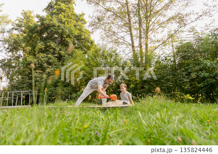 A father interacts with his young child in a lush playground surrounded by trees. They are happily playing with sand and enjoying each other's company outdoors on a bright day A father interacts with his young child in a lush playground surrounded by trees. They are happily playing with sand and enjoying each other's company outdoors on a bright day 135824446