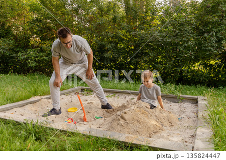 A father interacts joyfully with his child in a sandbox, sharing playful moments. Nature surrounds them, adding warmth to their bonding experience in a bright setting A father interacts joyfully with his child in a sandbox, sharing playful moments. Nature surrounds them, adding warmth to their bonding experience in a bright setting 135824447