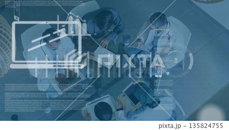 Collaborating team leaning over charts around glass table in office, with laptops and stethoscopes 135824755