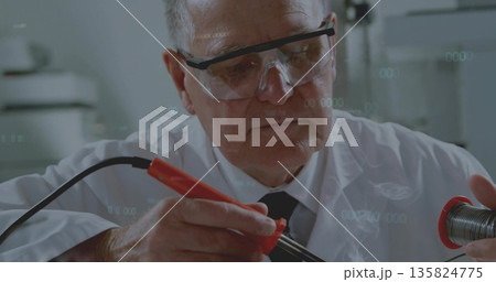 Using red-handled iron technician in white coat and goggles melting solder on wire at lab bench 135824775