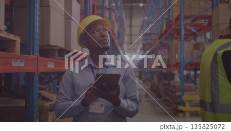 Staff inspecting stock holding tablet in warehouse aisle wearing safety gear among racks and boxes Staff inspecting stock holding tablet in warehouse aisle wearing safety gear among racks and boxes 135825072