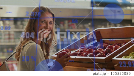Smiling woman wearing long-sleeve top holding red apple at supermarket bins with data overlays Smiling woman wearing long-sleeve top holding red apple at supermarket bins with data overlays 135825089