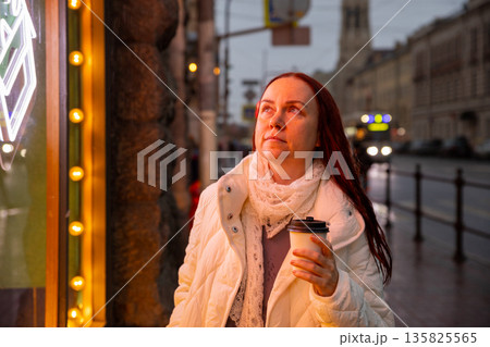 European woman with takeaway coffee stands dreamily under neon light with cup of aromatic coffee on street in evening European woman with takeaway coffee stands dreamily under neon light with cup of aromatic coffee on street in evening 135825565