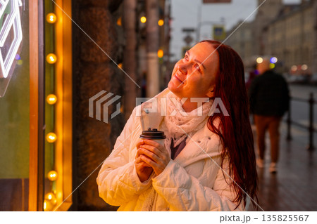 A happy woman drinks coffee in the evening on a neon lit street. A magical moment. A happy woman drinks coffee in the evening on a neon lit street. A magical moment. 135825567
