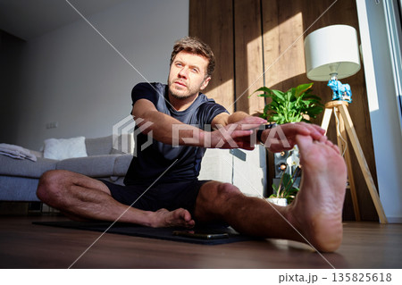 Man stretching legs while sitting on yoga mat at home in living room. Concept of home workout, flexibility training, daily workout and healthy lifestyle Man stretching legs while sitting on yoga mat at home in living room. Concept of home workout, flexibility training, daily workout and healthy lifestyle 135825618