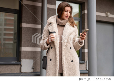 A Caucasian woman in a coat surfs the news on her smartphone and drinks coffee near the entrance to a residential building. Female uses smart phone for checking latest news, surfs sites. Businesswoman 135825790