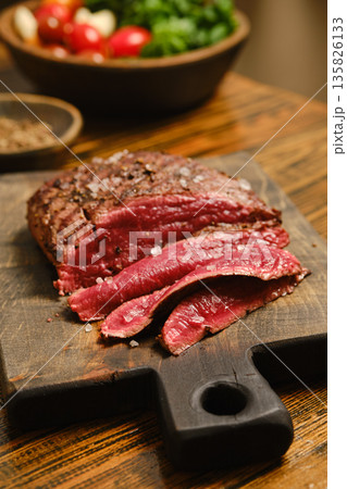 Sliced beef resting on wooden board with seasoning and fresh vegetables nearby 135826133