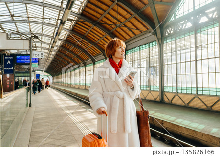 Caucasian mature woman with red hair standing on railway platform, holding smartphone and suitcase, waiting for regional train. Real travel, business trip, public transport, winter city Europe. Caucasian mature woman with red hair standing on railway platform, holding smartphone and suitcase, waiting for regional train. Real travel, business trip, public transport, winter city Europe. 135826166