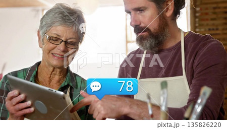 Pointing instructor in apron, mentee examining tablet showing 78 in studio, with brushes 135826220