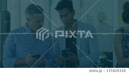 Two male coworkers comparing phone screens in office by window with sticky notes on glass partition 135826326