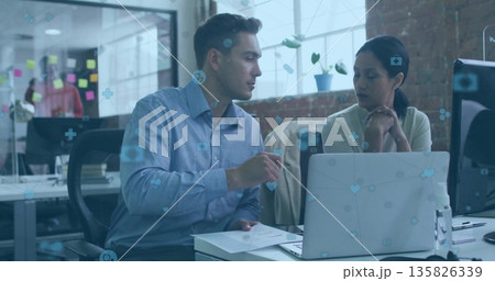 Two office colleagues pointing at laptop screen and watching in open-plan office, with sticky notes 135826339