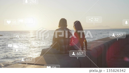 Sitting couple wearing casual clothes watching sunset on seawall facing ocean, with overlay icons 135826342