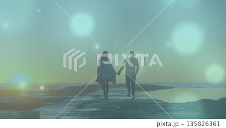 Walking couple holding hands on concrete pier over calm sea at dusk, poncho under lens flares Walking couple holding hands on concrete pier over calm sea at dusk, poncho under lens flares 135826361