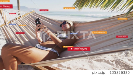 Reclining woman wearing swimsuit, sunglasses and headband, holding smartphone in hammock at beach Reclining woman wearing swimsuit, sunglasses and headband, holding smartphone in hammock at beach 135826380