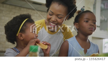Leaning mother and daughters eating pancakes at kitchen counter with syrup bottle and pancake stack Leaning mother and daughters eating pancakes at kitchen counter with syrup bottle and pancake stack 135826417