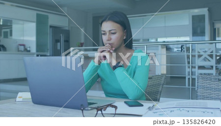 Typing remote worker wearing teal top, stud earrings in home kitchen, with laptop and smartphone Typing remote worker wearing teal top, stud earrings in home kitchen, with laptop and smartphone 135826420