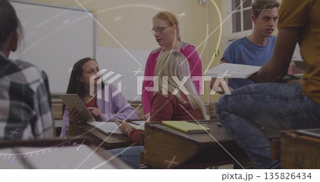 Collaborating students pointing at tablet screen in classroom, passing papers at wooden desks Collaborating students pointing at tablet screen in classroom, passing papers at wooden desks 135826434
