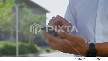 Tapping smartphone, man in white shirt and black smartwatch standing near parking, blurred trees 135826487