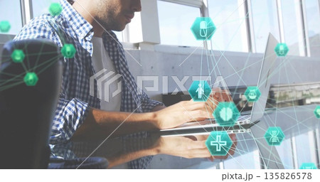 Typing Hispanic man in checkered shirt at glass desk, with silver laptop and floating digital icons 135826578