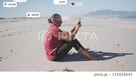 Capturing mature man streaming on sandy beach, holding smartphone showing overlay icons, copy space Capturing mature man streaming on sandy beach, holding smartphone showing overlay icons, copy space 135826645