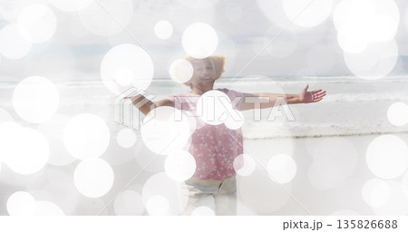 Senior woman in floral blouse stretching arms wide on shore by ocean waves, with bokeh overlay Senior woman in floral blouse stretching arms wide on shore by ocean waves, with bokeh overlay 135826688