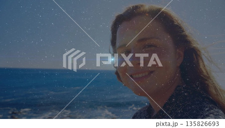 Standing woman in geometric shirt smiling at camera on beach edge, sunlight reflecting off water 135826693