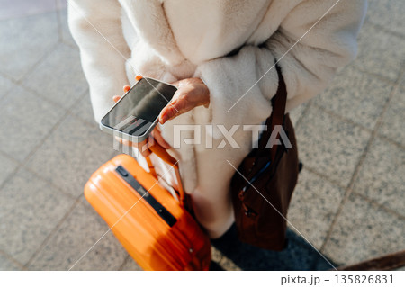 Close-up of female hand holding smartphone above orange suitcase handle, white faux fur coat and leather bag floor background, travel planning, digital navigation, modern transport routine Close-up of female hand holding smartphone above orange suitcase handle, white faux fur coat and leather bag floor background, travel planning, digital navigation, modern transport routine 135826831