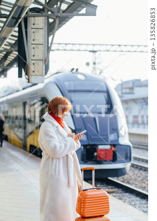 Middle aged Caucasian woman checking smartphone while standing near train, stacked orange suitcases on railway platform. Concept of ecology, work travel, business, everyday rail commuting in Europe. 135826853