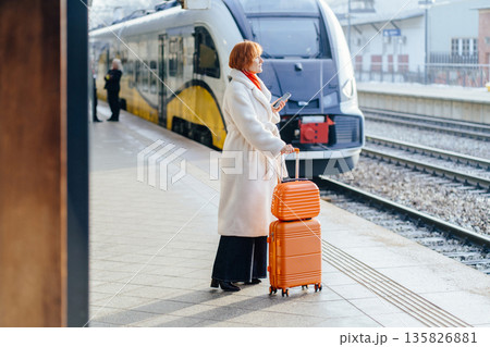 Mature Caucasian woman standing on railway platform beside orange suitcase, holding smartphone and looking toward arriving electric train. Professional routine, public transit, winter city journey. 135826881