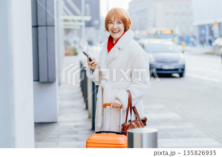 Smiling red-haired adult woman with suitcase uses smartphone while moving through modern city street. Solo travel, urban mobility, confident lifestyle, relocation moment, everyday digital navigation. 135826935
