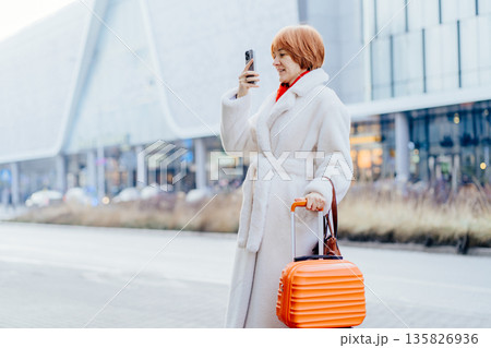 Thoughtful red-haired woman stands in city environment holding phone and suitcase. Urban travel pause, navigation moment, relocation process, modern mobility, independent everyday movement. 135826936