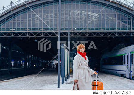 Mature woman waiting with suitcase on railway station platform. Concept of urban transit space, relocation moment, winter travel routine, mobility, public infrastructure, personal journey. 135827104