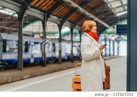 Mature woman waiting with suitcase on railway station platform near train. Concept of urban transit, relocation, public transport, winter travel, mobility, everyday movement, departure, journey. 135827202