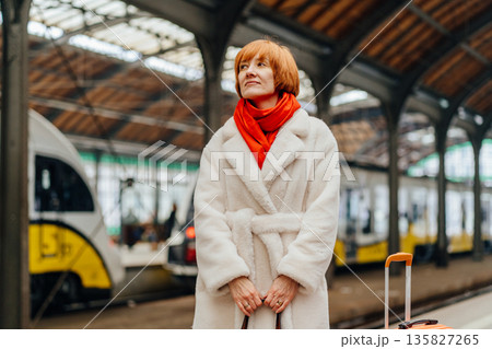 Mature woman in white faux fur coat and red scarf standing calmly on railway station platform. Concept of stability, self control, independence, inner balance, grounded mindset, slow living, mobility. Mature woman in white faux fur coat and red scarf standing calmly on railway station platform. Concept of stability, self control, independence, inner balance, grounded mindset, slow living, mobility. 135827265