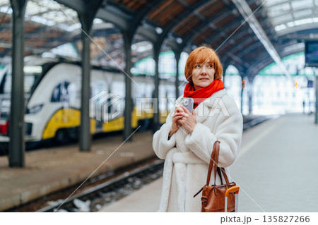 Portrait of mature red haired woman in faux fur coat and red scarf on railway station platform with suitcase. Concept of life transition, decision making,independence, new chapter, personal change. 135827266